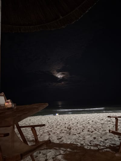 This nighttime photograph depicts a beach scene in Varna, Bulgaria. The upper section reveals a dark, thatched roof or umbrella. Beneath this, a dark night sky is visible, featuring a moon partially obscured by diffuse clouds, particularly on the left. Faint stars are discernable in the darker sky areas. The horizon line separates the sky from the ocean, where multiple lines of white foamy waves are breaking near the shore, reflecting available light. The lower half of the image consists of light-colored sand, exhibiting numerous footprints and cast shadows, suggesting illumination from an external source not fully in frame. In the lower-left foreground, an angled wooden table supports a small, glowing object, likely a candle or an ashtray with a light source. A partially visible wooden beach chair with a light-colored fabric seat is situated below the table. On the right side of the beach, near the water's edge, a small, light-colored object, possibly a cooler or bag, is present.