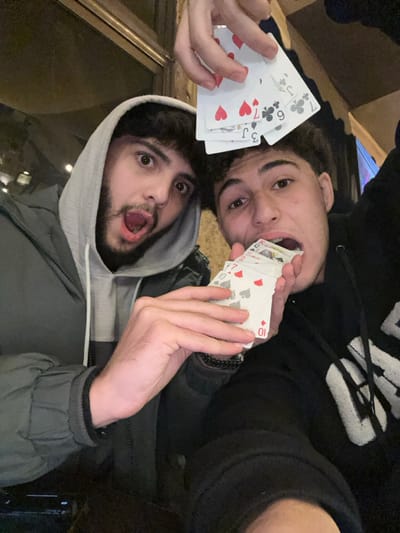 Two young men pose closely, holding playing cards. One, wearing a grey hooded jacket, opens his mouth in a surprised expression. The other, in a black hoodie, has cards in his mouth. The visible cards include a 7 of hearts, 6 of clubs, and Jack of hearts in one hand, and a 10 of hearts and 10 of spades in the other. The background appears to be dimly lit, possibly indoors.