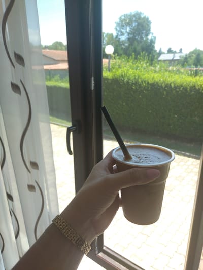 A hand with a gold bracelet and pink nail polish holds a to-go cup of coffee with a black straw. The cup is clear plastic and filled with a frothy brown beverage. In the background, through a window with a dark frame, a lush green hedge and a light-colored paved area are visible. A white curtain with brown abstract designs is on the left side of the frame. The scene appears to be outdoors, possibly in a residential area, given the presence of a hedge and what looks like a roofline in the distance. The lighting suggests a bright, sunny day. This image was taken in Yagoda, Bulgaria.