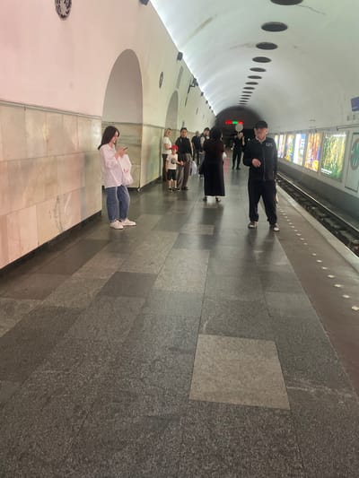 This image captures a scene at the Rustaveli metro station in Tbilisi, Georgia. A woman in a white shirt and blue jeans stands on the platform, looking at her phone and holding a white bag. To her right, a young boy in a red cap and dark shorts stands next to a woman wearing a black dress. Further down the platform, a man in a black jacket and pants is visible, facing the camera. The station features a curved, illuminated ceiling with circular openings and a tiled wall. Posters are displayed along the wall to the right of the platform. The floor is tiled with large, dark grey granite slabs. The overall atmosphere suggests a typical day at a public transportation hub.