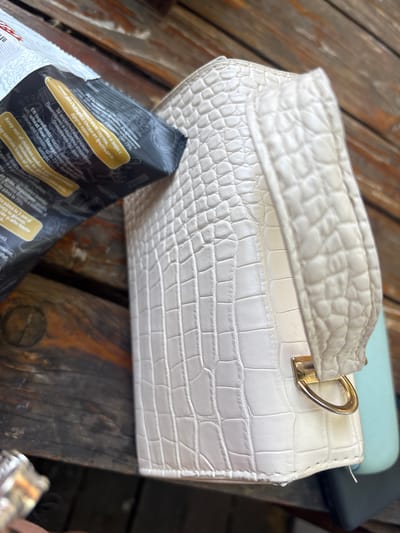 This close-up, eye-level shot focuses on a white, crocodile-embossed handbag resting on a rustic wooden surface, possibly a table or deck. The handbag has a structured shape with a handle and a gold-toned clasp. A portion of a dark, possibly black, package with visible text in multiple languages, including English, is in the foreground, partially obscuring the handbag. The text on the package is partially legible, with phrases like "EN Ingredients: roasted sunflower seeds salt wheat flour" and "Keep out of children under 3 years". The wooden surface appears weathered, suggesting an outdoor setting. The lighting suggests it is daytime. The scene is static, with no activity depicted beyond the placement of the objects. The location context provided is Nova Zagora, Bulgaria.