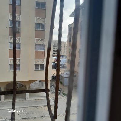 The image provides a view from an elevated position, looking out through multiple blurry, dark vertical metal bars. The scene depicts an urban landscape in Khan Yunis, Palestinian Territory, showing signs of extensive damage and disruption. On the left, a multi-story, light-colored residential building features decorative brown vertical sections and patterned window surrounds; numerous windows appear dark or damaged. The building displays visible structural damage, particularly at its base where arched openings lead to a recessed area containing debris, and on upper floors. Opposite this structure, an unpaved ground area is covered with dirt, dust, and rubble, including piles of fragmented concrete. In the central midground, a cluster of makeshift shelters, constructed from dark tarpaulins or fabric, is present, alongside smaller, simple concrete or metal structures. Further residential buildings, some also exhibiting damage, are visible in the background under an overcast sky. A similarly styled, multi-story building on the right is partially visible and shows significant damage to its upper sections, presenting a jagged, broken profile. No individuals or specific activities are discernible within the frame. A "Galaxy A14" watermark is visible in the lower-left corner.