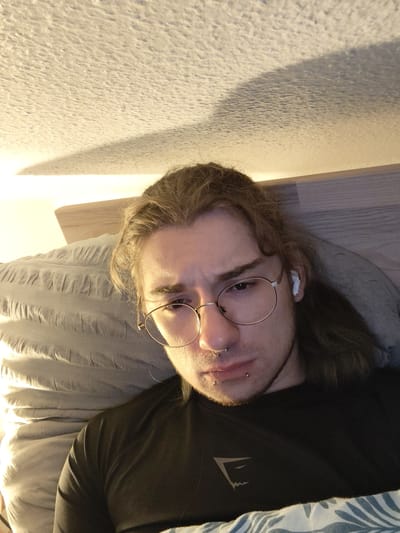 A close-up, overhead shot shows a young person with long, light brown hair, wearing round, gold-rimmed glasses and a black t-shirt with a white geometric logo. They are lying down on a bed with a grey, wrinkled duvet. The person has several facial piercings, including a septum ring and two lower lip rings. One wireless earbud is visible in their right ear. The background shows a textured ceiling with a shadow cast across it. The scene appears to be indoors, in a bedroom.