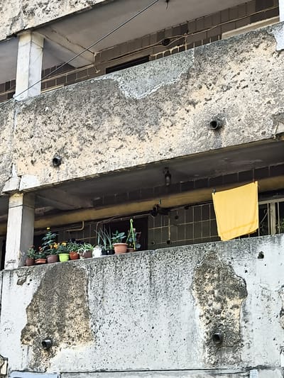 The image shows a close-up view of a concrete balcony of an apartment building in Donetsk, Ukraine. The balcony is cluttered with potted plants, some of which are flowering. A yellow towel is hanging on a clothesline stretched across the balcony. The concrete structure appears weathered and shows signs of wear and tear, with some areas of discoloration and damage. The background reveals the interior of the apartment through a window. The scene depicts an everyday urban setting, likely in a residential area.