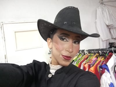 A person wearing a black cowboy hat and a black jacket with a white lace collar is posing for a selfie. The person has theatrical makeup on, including dark eyeshadow and red lipstick. Behind them, there is a rack of colorful costumes and a white door. The scene appears to be in a backstage area, possibly a dressing room, located in Porlamar, Venezuela.