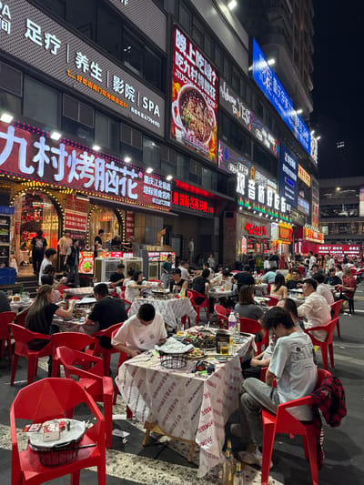 This vertical shot captures a bustling night market street scene in Liangjiang, China, filled with people dining outdoors. Numerous patrons, predominantly young adults, are seated closely at small tables arranged along the sidewalk, all using bright red plastic chairs. The tables are covered with white paper tablecloths, many displaying repetitive Chinese text and branding. Guests are actively eating from bowls, drinking from glasses and bottles, and socializing in a lively atmosphere. Empty bottles and discarded wrappers litter some tables, indicating ongoing meals.
The backdrop consists of multi-story buildings illuminated by numerous vibrant neon signs in various colors – blue, red, orange, and white – casting a colorful glow over the street. Visible text on the signs includes:
* A prominent blue sign for "重庆同善医院" (Chongqing Tongshan Hospital) with a phone number "023-68883396".
* A large red sign on a building reads "九村烤脑花" (Jiucun Roast Brain Flower), with "JIUCUN ROAST NOW" in English below it.
* Tablecloths feature text like "重庆非遗美食名片" (Chongqing Intangible Cultural Heritage Food Card), "九村烤脑花" (Jiucun Roast Brain Flower), and "JIUCUN ROAST NOW".
* Another long sign advertises services: "足浴 | 养生 | 影院 | SPA" (Foot Massage | Wellness | Cinema | SPA) with contact "联络电话 67799490".
* Other signs display "Welcome to YES!", "鲜虾", "老火锅", "红花蟹" (Red Flower Crab), and "重庆工行" (Chongqing Industrial and Commercial Bank).
Portable air coolers are visible between some dining groups. The scene suggests a popular street food destination, bustling with activity into the night.