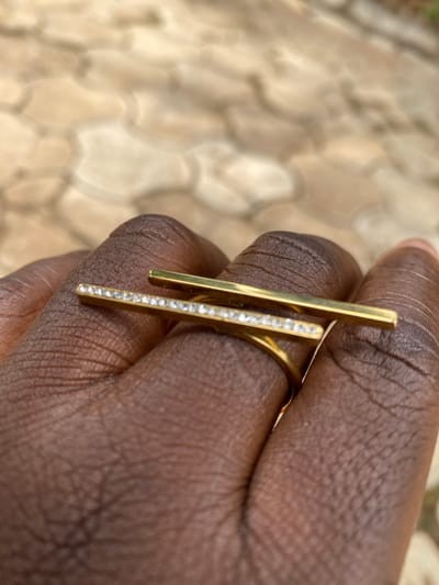 A close-up captures a person's brown hand, adorned with a gold-colored ring. The ring features a horizontal bar on top and a vertical bar accented with small, bright stones. The background is an out-of-focus pattern of light tan and brown shapes. The location is assumed to be Maigizo, Nigeria.