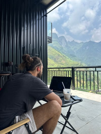 A man with long hair tied in a bun sits on a chair outdoors, working on a laptop. A drink with a straw sits on a table next to the laptop. In the background is a mountainous landscape, likely in Mèo Vạc Commune, Vietnam. The man is wearing a gray shirt and light-colored shorts. The scene appears to be a balcony or terrace with a black metal railing and a black corrugated metal wall.