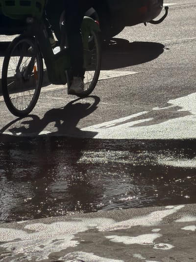 The media file captures a high-contrast, sunlit scene from an overhead or slightly elevated perspective, rotated clockwise. The ground, likely pavement or asphalt, is divided into intensely lit and deeply shadowed areas. On the left, a bicycle is partially visible, showing its front wheel, part of the frame, and a shoe resting near a pedal.
Prominently featured are long, dark shadows cast across the bright ground. The most distinct shadow belongs to a person on a bicycle, stretching from the unseen rider to the right. Other elongated, irregular shadows also extend across the pavement, suggesting the presence of additional objects or structures outside the frame.
To the right of the paved area, a dark, reflective surface, likely a puddle or stream of water, catches the strong sunlight. This water body is covered with numerous sparkling points of light, indicating a shimmering or rippled surface under direct sun. The bright areas of the pavement also exhibit a shimmering, speckled texture due to the intense light.
The strong, low-angle shadows and bright reflections suggest either early morning or late afternoon on a clear, sunny day. The scene conveys an urban setting consistent with the provided location context of Paris, France. No discernible text is visible in the image.