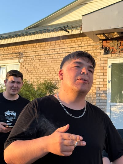 A photograph shows two men in an outdoor setting. The main subject in the foreground is holding a lit cigarette, from which smoke is emanating, and has a silver chain around his neck. He is wearing a black t-shirt. Partially visible behind him and to the left is another man, also wearing a black t-shirt with a white Stussy logo. In the background, there is a brick building with a window. To the left of the brick building is a light yellow structure. The location is potentially in the city of Кемуль, Russia.
