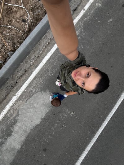 The image is a high-angle selfie taken outdoors, showing two subjects on an asphalt surface marked with white lines. One subject, in the foreground and center, is a person looking up at the camera, wearing a camouflage-patterned shirt. Their face is visible, and they appear to be taking the photo with their right arm outstretched.
To the left and slightly behind the first subject is a young child, facing away from the camera and looking downwards. The child is wearing a blue shirt with a graphic design and dark pants.
The scene is a paved area, possibly a road or walkway, with a concrete curb and dirt/gravel on the right side. The lighting suggests it is daytime, but the weather is overcast or diffused. There is no visible text in the image. The overall impression is of a casual outdoor moment between an adult and a child.