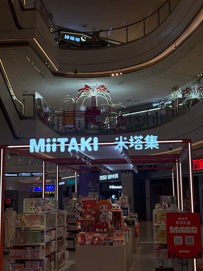 This is an interior shot of a store called "MiiTAKI" (米塔集), and the Chinese name above it translates to "stick." The store is located in the Liangjiang district of an unidentified city in China, probably in a shopping mall. The store sells merchandise, including figurines and collectibles. The image captures various details within and around the store: displays of Hisense products, store signage with the name "MiiTAKI" in both English and Chinese, a promotional sign indicating a gift of 100 yuan for new members, and other stores like "Nikoko" and "Weivilia." The environment includes the interior architecture of the shopping mall with lighting and decorative elements. No people are directly visible in the frame, but the presence of merchandise and signs indicates the store is open for business.