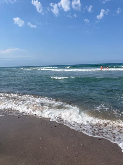 A vertical, wide-angle shot captures a serene beach scene at Ahtopol, Bulgaria. The ocean, a dominant feature, displays a vibrant turquoise hue under a clear blue sky dotted with fluffy white clouds. Gentle waves roll towards the shore, creating a foamy white surf that laps at the dark, sandy beach. In the distance, three individuals are visible wading in the shallow water, suggesting a leisurely day at the beach. The sun appears to be high in the sky, indicating midday, and the overall atmosphere is one of tranquility and relaxation. The composition emphasizes the vastness of the sea and sky, with the shoreline acting as a clear demarcation between the water and the land. No text is visible in the image.