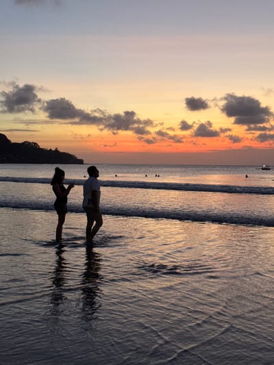 The image depicts a serene sunset scene at a beach in Kuta Selatan, Indonesia. The sky is a gradient of soft hues, transitioning from a pale lilac on the left to warm oranges and yellows as it nears the horizon, with scattered dark clouds adding texture. Silhouetted against this vibrant sky are two people standing in the shallow water. A man, wearing a light-colored short-sleeved shirt and shorts, faces a woman who is looking down at her phone. The woman is dressed in dark clothing and her hair is tied up. Their reflections are clearly visible on the wet sand and rippling water, creating a sense of symmetry and calm. Gentle waves are breaking on the shore, their foam creating delicate patterns on the wet sand. In the distance, the ocean stretches out, with a few small boats visible on the horizon, hinting at a calm sea. A dark, silhouetted landmass, possibly a hill or a section of coastline, is visible on the far left. The overall mood is peaceful and romantic, evoking a sense of quiet enjoyment of the natural beauty of the sunset. The image appears to have been taken during the golden hour, just before sunset. No text is visible in the image.