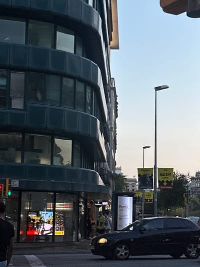 A dark-colored hatchback car is in the foreground, driving on a city street in Barcelona, Spain. The car is moving from left to right in the frame, and its headlights are on. To the left of the car, a large building with a dark facade and multiple windows dominates the scene. Several streetlights and posters are visible in the background, some of which advertise events or artists. The sky is clear and a light blue, suggesting it is either early morning or late afternoon.