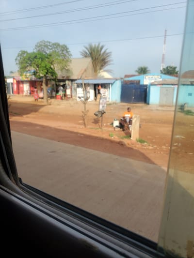 The image depicts a street scene in Kakau, Nigeria. In the foreground, a portion of a vehicle window is visible, framing the view of the surroundings. The street itself is made of dirt and gravel, with a paved road running through it. Several small businesses and residential structures line the street, some painted in bright colors. A few people are visible, including a person in an orange shirt sitting near a post and a woman in a patterned dress walking in front of a building. There are also trees and a telecommunications tower in the distance. The overall impression is that of a typical day in a bustling Nigerian town.