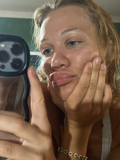 This is a close-up, vertically oriented, selfie-style photograph of a young person. The person has fair skin, blonde hair, and blue eyes. Their face is turned towards the camera, and one hand is held up to their cheek, partially obscuring their face. Their lips are slightly parted. The person is wearing a white top. A bracelet with small, flower-like charms is visible on their right wrist. The background is out of focus but appears to be an indoor setting with a green wall and some reflections, possibly from a mirror or phone screen. The lighting suggests it could be daytime or a well-lit indoor environment. There is no visible text in the image. The overall impression is casual and personal.