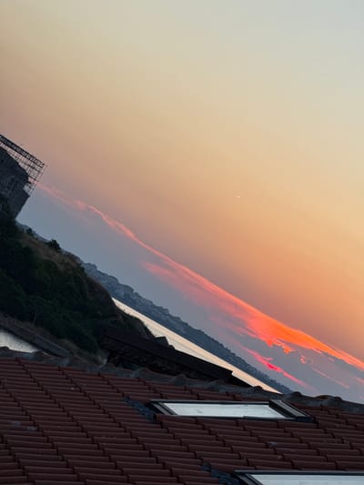 This is a digitally captured media file, likely a photograph, depicting a twilight scene with a prominent building in the foreground. The building, constructed with reddish-brown tiles, features at least two windows with dark frames. The scene is oriented such that the horizon line runs diagonally across the frame, from the lower left to the upper right.
In the distance, a body of water, likely the sea, reflects the soft light of the sky. Along the coastline, there are hints of buildings or structures nestled into the landscape, and dense green vegetation covers parts of the elevated terrain. A dramatic sky dominates the upper portion of the image, with wisps of clouds catching the last rays of the sun, painted in hues of orange, red, and pink. A faint crescent moon is visible in the pale orange sky. In the lower left, a large, angular structure resembling scaffolding or a construction framework is partially visible.
The overall mood is serene and somewhat dramatic due to the vibrant sky. The time of day is clearly sunset or twilight, indicated by the colors of the sky and the quality of light. The location context suggests Sozopol, Bulgaria, which would place this scene along the Black Sea coast. There is no visible text in the image.