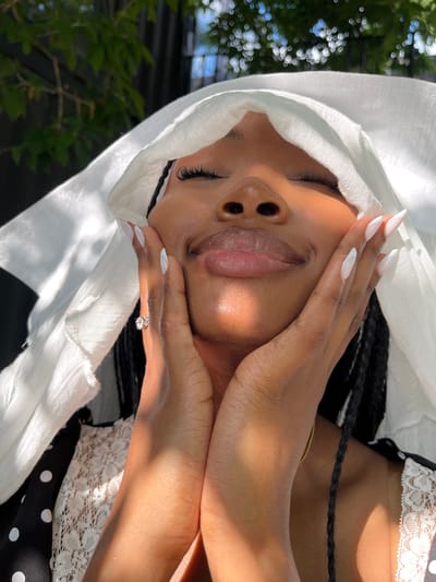 An upward-angle, close-up shot shows a young Black woman with her eyes closed and a gentle smile. She is looking up towards a bright light, which casts dramatic shadows across her face and chest. She is wearing a white headscarf that partially covers her hair, which is styled in long braids. Her hands are placed on her cheeks, with her fingers extending towards her forehead. She is wearing a black dress with white polka dots and a white lace trim. A ring with a diamond is visible on her left ring finger. The background is blurred and appears to be outdoors, with trees and a fence visible. The image is well-lit, with the sun shining brightly.