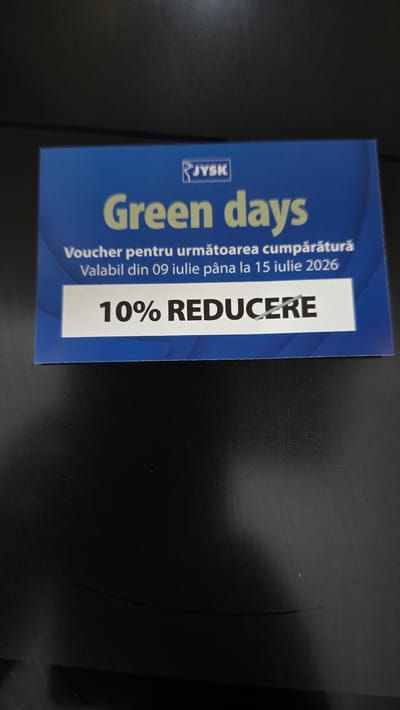 The image shows a close-up of a JYSK voucher. The voucher has a blue background with white text and a graphic element. The text reads "Green days" in large font. Below this, it says "Voucher pentru următoarea cumpărătură" which translates to "Voucher for the next purchase." Further down, it states "Valabil din 09 iulie până la 15 iulie 2026," meaning "Valid from July 9 to July 15, 2026." At the bottom, in a white rectangular box, is the text "10% REDUCERE," indicating "10% DISCOUNT." The voucher appears to be on a dark, textured surface, possibly a table or desk. There is no indication of any specific location in Chișinău, Moldova, nor are there any people or actions depicted.