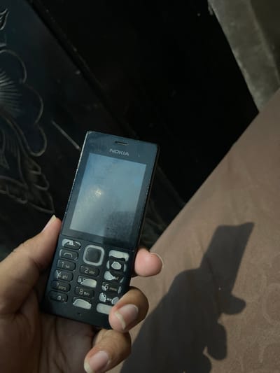 A close-up, eye-level shot shows a person's hand holding a black Nokia feature phone. The phone's screen is visible and appears dirty, with a faint reflection. The keypad is clearly visible, with numbers and letters arranged in the standard layout. The background is dark and out of focus, with a hint of a patterned surface on the left and a textured brown fabric on the right, casting a shadow of the phone and hand. The overall lighting suggests an indoor setting. There is no identifiable information about the location within the city of Bandarbireuen, Indonesia.