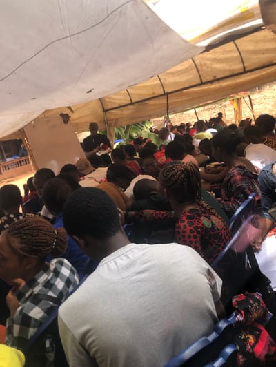 The image captures a densely packed outdoor gathering within Akpaka Forest Reser, Nigeria, beneath a light-colored, tarp-like canopy supported by a metal frame. Numerous individuals, predominantly adults of African descent, are seated in rows, facing towards the front of the tent. In the mid-ground, a male figure wearing a dark shirt and cap is positioned behind a white-draped table, holding a red object in his right hand while appearing to address the assembly. A whiteboard with illegible text stands to his left. Attendees are dressed in various casual attire, with some individuals exhibiting braided hairstyles. Visible objects include a patterned bag on the table, blue plastic chairs, and scattered papers. The environment beyond the tent structure includes a light-brown earthen wall and green foliage. Strong overhead sunlight illuminates the scene, casting shadows.