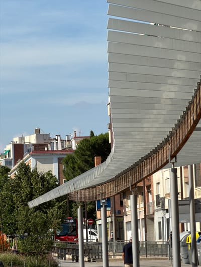 The image shows a street scene in Terrassa, Spain, focusing on a modern architectural structure with a curved roof made of metal louvers supported by wooden beams. In the background, a red bus with "VILLADA" displayed on its front is visible. Several buildings with multi-story facades and balconies line the street. Green trees and foliage are scattered throughout the scene. The sky is a pale blue with scattered clouds. The foreground features a metal railing and some street furniture, including benches. There are a few individuals in the scene, including a person walking away from the camera and another sitting on a bench. Trash receptacles are also present. The lighting suggests it is daytime, with the sun casting shadows.