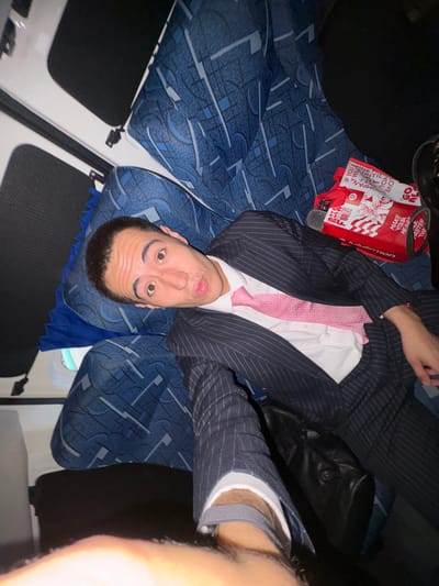 A man in a pinstripe suit and pink tie is lying in a bus seat. The bus has blue and white patterned upholstery. A red Lululemon tote bag sits next to him. He is taking a selfie, and part of his bare leg is visible. The bus window and interior are visible. The location is assumed to be in Mexico City, Mexico.
