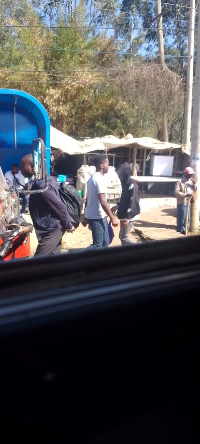 The image depicts a roadside scene in Ngong ward, Kenya, under clear daylight conditions. In the immediate foreground, a dark vehicle interior frame partially obscures the lower portion of the view. The mid-ground features a blue-colored vehicle, possibly a tuk-tuk or small utility truck, positioned on the left. A male subject, dressed in a dark jacket and black trousers, is partially visible from the back, holding a black backpack, standing adjacent to the blue vehicle. Another male subject with a bald head and white t-shirt is seated further left within or near the vehicle. To the right, two young male subjects are observed in close proximity; one wears a white t-shirt and blue jeans, holding a purple bag, while the other wears a black hooded jacket. Further right, a male subject in a light-colored shirt and hat, holding a wooden stick, stands near a light-colored utility pole. The ground surface consists of unpaved, exposed earth, with visible wooden planks forming a section of a path. In the background, several makeshift market stalls with white tarpaulin or corrugated metal roofs are present, one displaying a white refrigerator. Beyond these structures, dense green and dry vegetation, including tall trees and utility lines, are visible against a clear blue sky.