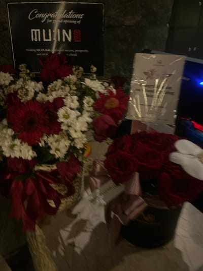 In the city of North Kuta, Indonesia, a close-up shot captures a celebratory scene featuring floral arrangements and a congratulatory sign. The focal point is a large basket overflowing with a mix of red gerbera daisies, white chrysanthemums, and other red and white flowers, tied with a prominent red ribbon. To the right, a round black hatbox arrangement of deep red roses is visible, adorned with a delicate pink ribbon. Behind the flowers, a black rectangular sign displays white text that reads "Congratulations for grand opening of MUIN Wishing MUIN Bali continued success, prosperity, and an inventory ahead." Partially obscured by the flowers, a white card or plaque with a floral motif is also present. The background is dark and indistinct, suggesting an indoor setting or nighttime environment.