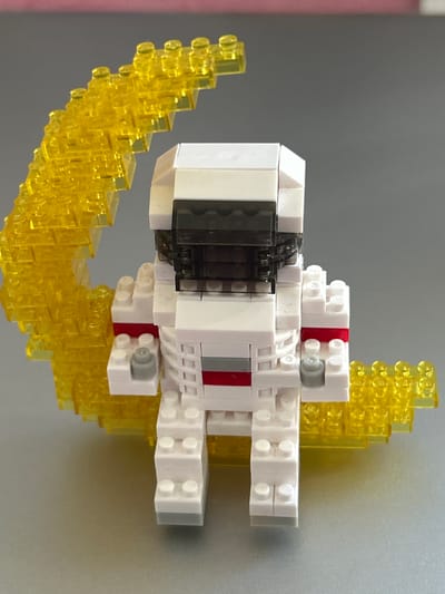 The image depicts a Lego model of a spacecraft with yellow exhaust trails, against a grey background with a hint of pink on the left. The spacecraft is primarily white with some black, grey, and red details. The yellow exhaust trails are made of translucent yellow Lego bricks. No people or location in Strasbourg, France, are visible in the image.