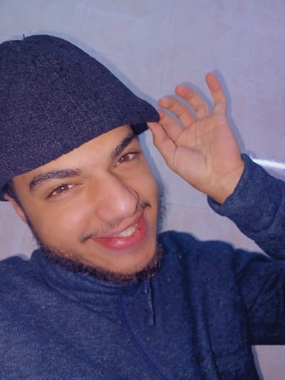 A person with dark hair and a beard is lying on their back, smiling. They're wearing a dark gray hooded sweatshirt, with the hood pulled partially over their head. One hand is raised, fingers slightly curved, near the edge of the hood. The background appears to be a plain, light-colored surface. The image is rotated, giving a tilted perspective.