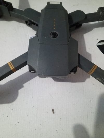 A gray drone sits on a textured white surface. The body of the drone is in the center, featuring a power button and battery indicator lights. Two arms with gold stripes extend towards the upper left and lower right corners. The shadows suggest the light source is above and slightly to the right. Debris is scattered on the white surface, including a dark speck near the bottom center.
