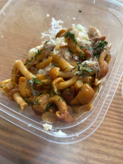 A close-up, eye-level shot shows a clear plastic container filled with pickled mushrooms, garnished with dill. The mushrooms are a rich, glossy brown and are mixed with a creamy white dressing. The container is sitting on a wooden table. The image is taken in Kaliningrad, Russia.