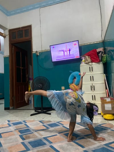 This media file shows a young girl doing yoga in a room. She is wearing a light blue t-shirt with a cartoon princess printed on it and a light blue skirt. She has a purple bow in her hair. She is attempting a yoga pose while following an exercise video on a television screen. The television displays a yoga instruction video with cartoon figures demonstrating poses. Beside her, a blue neck pillow with a burger toy is on the floor. A black oscillating fan is behind her. The room appears to be a living room or playroom, with a patterned rug on the floor and a wooden door in the background. There are also cabinets and some bags visible in the room, suggesting a home environment. The image is taken in Gelam, Indonesia. The lighting suggests it could be daytime.