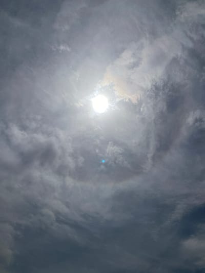 The image depicts a bright sun high in a cloudy sky above Barden Yana, Nigeria. The sun, a brilliant white orb, is positioned in the upper center of the frame, partially obscured by thin, wispy clouds that create a bright glare. Surrounding the sun, a distinct, faint circular solar halo, characterized by a subtle rainbow effect, is visible against the darker, predominantly gray and white scattered clouds that fill the rest of the sky. A small, prominent blue lens flare is present directly below the sun's position. No other subjects, objects, people, actions, or interactions are discernible; the scene is entirely an outdoor sky view.