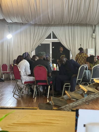 The image depicts an indoor gathering, likely a social event or meeting, taking place in what appears to be a large hall or venue. The setting is decorated with flowing drapes that create a soft, diffused lighting effect. In the foreground, a group of people are seated around tables, engaged in conversation. Their attire suggests a formal or semi-formal occasion, with some individuals wearing traditional Nigerian clothing, including caps.
The people are of various ages and genders. Some are looking at each other, while others are looking down or away. The atmosphere seems relaxed, with some individuals appearing deep in thought or conversation. There are several chairs, tables, and what seem to be decorative elements or service items on the tables. The floor is made of polished wood, reflecting the ambient light. The background features a large window with curtains and additional lighting fixtures. The overall impression is one of a lively social gathering in a well-lit indoor space.
Specific to the location in Barakin Lerawi, Nigeria, the traditional attire worn by some individuals strongly indicates a Nigerian context. The style of the venue and the social gathering aligns with typical events held in Nigeria. However, without more specific visual cues or identifying landmarks within the image itself, it's impossible to pinpoint an exact building or precise location within Barakin Lerawi. The scene is characteristic of community events, celebrations, or formal meetings common in Nigerian cities and towns.
