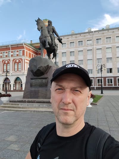 A selfie captures a middle-aged Caucasian man wearing a black baseball cap and a black t-shirt, standing in a public square. He is positioned in the foreground, with a monument of a man on a horse behind him. The monument is on a stone pedestal with an inscription. In the background, there are two buildings: one is a light-colored building with many windows, and the other is a red building with arched windows. The square is paved with grey cobblestones, and there are some potted plants and streetlights visible. The sky is blue with some clouds. The location is the National Congress of the Republic of Mari El in Yoshkar-Ola, Russia, identified by the iconic Monument to Ivan the Terrible, commissioned by Ivan IV's father, Vasily III.