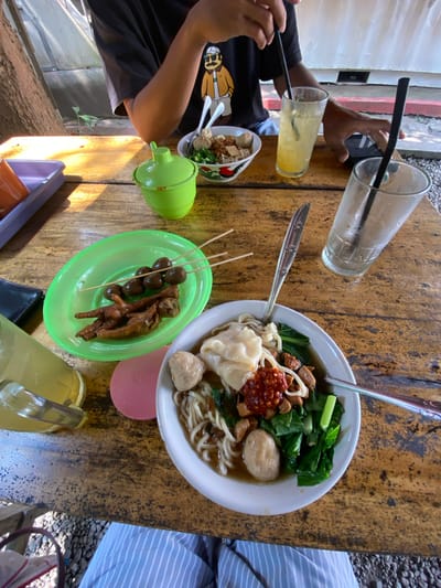 This media file captures a top-down view of a casual meal set on a rustic wooden table, likely at an outdoor or semi-outdoor eatery in Madiun, Indonesia.

The primary subject is a large bowl of *Mie Ayam Bakso* (chicken noodles with meatballs) served in a white bowl, positioned slightly to the right of the center. It contains noodles, dark brown chicken pieces, green leafy vegetables (possibly bok choy), two large meatballs, and a *wonton* or *pangsit* dumpling, topped with a dollop of red chili paste. To the left of the main bowl, a bright green oval plate holds skewers of *sate ceker* (chicken feet) and *sate telur puyuh* (quail eggs), indicating common Indonesian street food snacks. A smaller bowl of noodles is also visible on the far left.

Beverages include a clear glass with a yellow/orange drink and a black straw, held by a person on the left whose hands and part of a black t-shirt with a yellow graphic are visible. Another empty clear glass with a black straw is near the top center. A yellow jug or pitcher is seen in the bottom right corner.

Two individuals are partially visible. On the left, a person is holding a drink and appears to be interacting with a phone. On the right, only a leg wearing light blue and white striped pants is visible.

The wooden table has a worn, light brown appearance. Various items are scattered across it, including spoons, a green lidded condiment container, a black wallet, and a pink round coaster with the word "Marin" faintly visible. The background shows a light-colored wall with red accents on the left and a gravel ground area on the right, suggesting a bright, sunny daytime setting. The scene evokes a relaxed, informal dining experience.