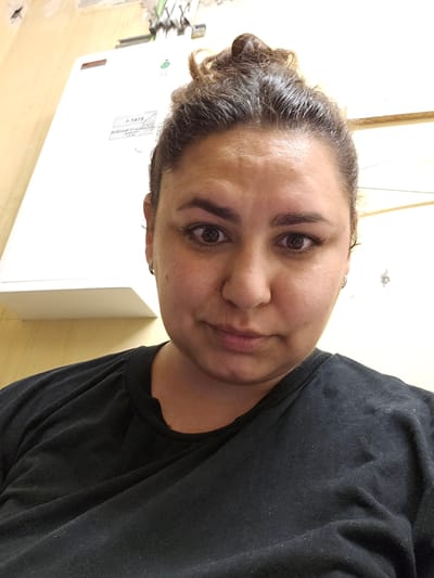A selfie of a woman with dark, curly hair pulled up into a bun is shown. She is wearing a black t-shirt. Her face is partially obscured by a white rectangular object in the background, which appears to be a piece of equipment with some labels. A beige wall is visible to the left, and a white wall to the right. The scene appears to be indoors, possibly in a utility area or workshop. The lighting is artificial. The image is shot from a close-up perspective, focusing on the woman's face and upper torso. The background suggests an industrial or commercial setting.