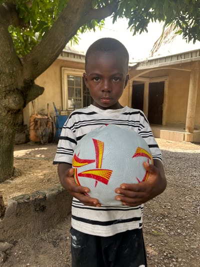 A young dark-skinned boy, approximately 5-8 years old, stands centrally in an outdoor setting within Mararaba, Nigeria. He has short dark hair and looks directly forward with a neutral expression. He is wearing a white t-shirt with horizontal black stripes and dark shorts. Both hands hold a white and grey soccer ball with red and yellow geometric patterns against his torso.

To the boy's left, a large tree trunk occupies the foreground, with its green foliage visible above. Behind the tree, a light-colored building with a visible window is present. Two wheelbarrows, one orange and one blue, are situated near the building. To the boy's right and extending into the background, another light-colored building features two dark doors and a raised entrance. A round pillar stands further right, supporting an overhang. The building's roof, appearing to be corrugated metal, is visible under bright sunlight. The ground is composed of packed dirt and small gravel or stones.