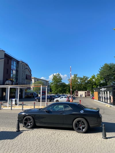 This media file depicts an outdoor scene in Minsk, Belarus, on a clear, sunny day. The primary subjects are several cars parked in a lot, with a black sports car prominently featured in the foreground. The setting appears to be a commercial or residential area, with buildings of various architectural styles visible in the background. Trees with lush green foliage line the area, suggesting a pleasant season, likely summer.
A boom barrier, red and white, is partially lowered, indicating a controlled entry or exit point for the parking area. A few people are visible near the barrier and in the background, some interacting with cars or walking. The time of day is clearly daytime, with strong sunlight casting distinct shadows on the paved ground. The overall mood is one of everyday activity in a developed urban environment.
While no significant text is clearly legible on the cars or buildings, there are signs on the buildings that are too small or blurry to be read. The focus remains on the vehicles, the parking infrastructure, and the surrounding environment under a bright blue sky.