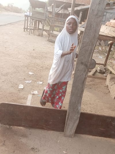 A young person, likely a girl, stands in the foreground, wearing a white hijab that covers her head and shoulders, and a patterned skirt with red, blue, and white geometric shapes. She has dark skin and is wearing dark sandals. Her mouth is slightly open, and she looks directly at the camera with her right hand raised and partially open, as if speaking or gesturing.
The setting appears to be a dirt-covered outdoor area, possibly a roadside or marketplace, consistent with Ebiya, Nigeria. The ground is unpaved and dusty, with scattered pieces of white litter, possibly paper or plastic. In the immediate foreground, a long, dark horizontal wooden plank lies on the ground, and a thick, rough-hewn vertical wooden beam stands upright to the right of the person, partially obscuring the scene. A circular, metallic object is attached to this vertical beam, with the text "MADISON" and "50 KG" faintly visible on its surface.
In the mid-ground and background, several simple wooden structures, resembling stalls or shelters, are visible. Some have corrugated metal roofs. A dirt road stretches into the distance on the left side of the image, where a figure, appearing to be an adult man in light clothing, can be seen further down the road. The lighting indicates it is daytime, with a warm, bright tone. The overall scene conveys a candid moment in a semi-urban or rural environment.