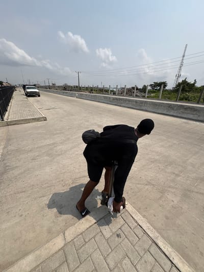 The image captures an outdoor scene in Igboefon, Nigeria, under a bright, partly cloudy sky. A dark-skinned individual, seen from behind and bending forward, is actively engaging with a metallic lifting device, possibly a hydraulic jack, positioned at the juncture of a light gray concrete roadway and a darker gray brick-paved sidewalk. The individual wears a black ribbed long-sleeved top, black shorts, black Nike slides on their feet, and a black head covering; a black cross-body bag is slung over their back. Their right hand, wrapped in a white cloth, grips the device's handle, which exhibits metallic disc-like components near its base.
The concrete roadway extends into the background, flanked on the left by a low concrete curb and a black metal fence bordering the sidewalk. A white pickup truck is discernible in the far distance on the left side of the road. To the right of the roadway, a concrete barrier delineates an area characterized by scattered construction materials, rudimentary fencing, sparse vegetation, and several utility poles supporting overhead power lines. A tall, lattice-structured communication tower is visible in the far background on the right. Strong shadows indicate direct sunlight.