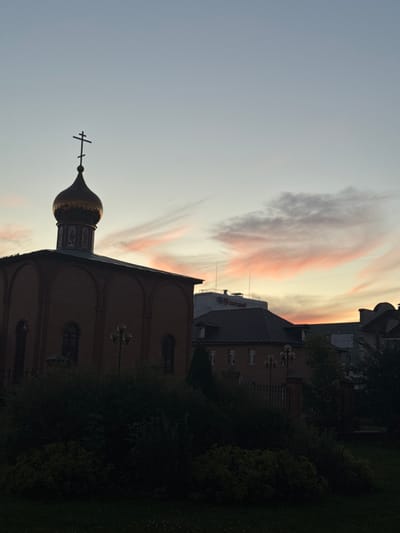 The image shows an Orthodox church with a golden dome and a cross on top, viewed from a low angle. The church is made of red brick and has arched windows. Behind the church, there are other buildings and trees. The sky is a pale blue with wispy pink and orange clouds, indicating it is either sunrise or sunset. The lighting suggests it is either early morning or late evening. The image appears to be taken in Shcherbinka, Russia, as indicated by the style of the church and its surroundings. There is no visible text in the image.