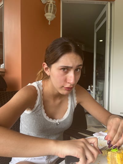 The image depicts a young female individual seated at a dark, possibly wicker, table, engaged in consuming corn on the cob. The scene is located in a residential outdoor or semi-outdoor space, such as a balcony or patio, in Floridablanca, Colombia.
The individual, appearing to be in her late teens or early twenties, has medium-brown hair pulled back with a visible bobby pin on the left side of her head. She wears a grey ribbed tank top with white lace trim along the neckline and armholes, featuring a small white bow at the center of the chest. Her skin appears untanned, and her brown eyes are directed towards the viewer with a neutral to slightly questioning expression. Both hands are visible; the right hand holds a black-handled knife, while the left hand holds a silver fork with a bright green, nubbly corn-on-the-cob holder attached. Her fingernails are painted with a dark, possibly plum-colored, polish.
On the table in front of her is a white plate containing a partially eaten yellow corn cob and scattered corn kernels. A crumpled white paper napkin rests on the plate.
The background includes an orange-brown wall on the left, to which a white, multi-tiered outdoor wall light fixture is mounted. To the right, an open white-framed door leads into a darker interior room. Within this room, reflective surfaces and various indistinct objects are visible. The lighting suggests a bright daytime setting.
