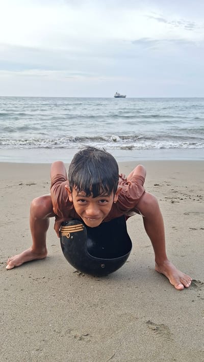 A young boy is on a sandy beach, positioned low to the ground, with his hands and feet supporting his weight. He is looking directly at the camera with a playful expression. In front of him, on the sand, is a black helmet with a gold stripe and the word "Classic" written on it. The ocean with gentle waves is behind him, and a large ship is visible in the distance on the horizon. The sky is overcast. The location is the beach in Lhokseumawe, Indonesia.