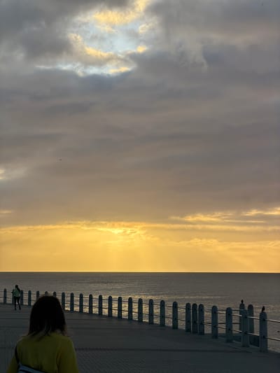 The image captures a scene at dusk or dawn in Cape Town, South Africa, along the coastline. The sky is a mix of gray and golden clouds, with the sun's light reflecting on the ocean. A concrete promenade runs along the coast, bordered by a series of evenly spaced concrete bollards connected by metal railings. A few people are present: one person wearing a yellow shirt is visible in the foreground, and two figures can be seen walking in the distance along the promenade. Two birds are perched on the railing near the horizon. The overall atmosphere is tranquil and serene, with a clear contrast between the city infrastructure and the natural environment.