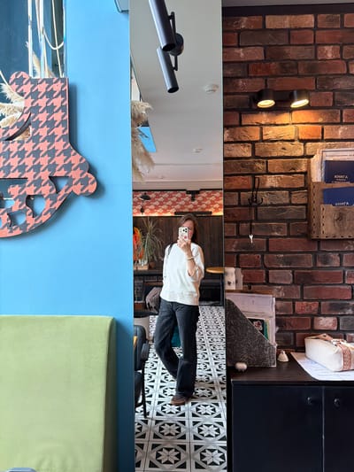 An interior shot depicts a woman taking a mirror selfie in a commercial establishment located in Izhevsk, Russia. The woman, with medium-length brown hair, wears a loose white long-sleeved top and dark jeans, and is holding a light-colored smartphone to capture her reflection. A light-colored bracelet is visible on her left wrist.
To her immediate left, a bright blue wall is visible, adorned with a large, abstract decorative element featuring a pinkish-orange and dark gray houndstooth pattern. Partially reflected in the mirror behind this decoration are dried ornamental grasses. A segment of a light green upholstered bench or seating unit is visible in the lower-left corner against the blue wall.
To her right, a prominent brick wall is featured, showcasing bricks in various shades of red, brown, and gray. Mounted on this wall are two black cylindrical spotlights emitting warm light downwards. Below these lights, a black three-pronged coat hook holds a small silver-colored key on its lowest peg. To the right of the hook, a wooden wall-mounted organizer contains several blue folders or books with white Cyrillic text, including "КНИГА" (Book) and "МИНПРОМТОРГ УР" (Ministry of Industry and Trade of Udmurt Republic), which identifies the location as being within the Udmurt Republic, with Izhevsk being its capital.
Below the organizer, a dark-colored counter or cabinet with two black handles supports various items: a textured dark brown magazine holder, a small white bell-shaped object, another small white irregular object, a stack of papers, and an elongated white fabric pouch with a striped orange and white handle.
The floor throughout the visible space features a geometric pattern of black, white, and gray tiles arranged in repeating star-like or compass rose motifs.
The mirror's reflection behind the woman reveals a patterned wallpaper with a repeating reddish-orange and light gray fish or cloud-like design, situated above dark wooden paneling. A tall green potted plant rests on a dark table, next to various small bottles. A black wall-mounted lamp is visible on the patterned wall in the reflection. The ceiling in the reflection is white, with a smoke detector or recessed light fixture visible.