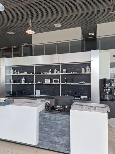 This is a photograph of the interior of a modern commercial space, possibly a dealership or a showroom, located in Izhevsk, Russia. The scene depicts a clean, well-lit environment with a prominent display shelf unit featuring a dark, patterned background with blue circuit-like lines. The shelves are organized with various decorative items and product displays, including white lanterns, a white mug, a white baseball cap, a plush toy dog, a black water bottle, and miniature car models. There is also a white sign with the text "#IAECO".
In the foreground, a white reception desk is visible, with a dark gray, marbled countertop. On the desk, a computer monitor, a printer, and some brochures are placed. To the right of the reception desk, a large, dark coffee machine is situated. The ceiling is a gray, tiled grid, with a prominent industrial-style pendant light fixture hanging down. The overall impression is one of a contemporary and professional setting. There are no people visible in the image.